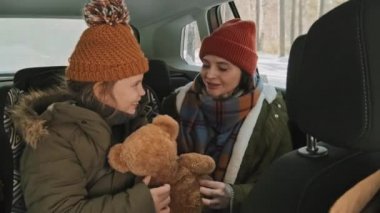 Yavaş çekimde, mutlu genç bir kadın ve 8 yaşındaki pelüş oyuncaklı bir kız arabanın arka koltuğunda oturmuş, kışın seyahat ederken oyun oynuyorlar.