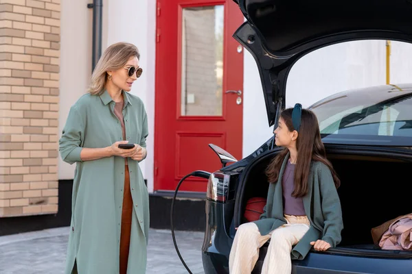 Schattig meisje zitten in de auto kofferbak en praten met haar moeder op laadstation — Stockfoto
