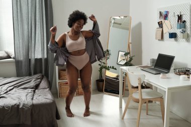 İç çamaşırlı, gri tişörtlü, yatak odasında dans eden neşeli genç bir kadın.