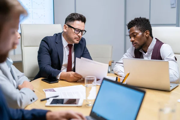 Fiatal komoly üzletember néz papírra, miközben érett kollégája elmagyarázza az adatokat — Stock Fotó