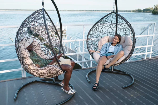 Zwei glückliche junge interkulturelle Freunde sitzen in Sesseln auf der Seebrücke — Stockfoto