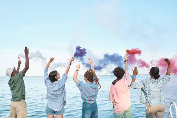 Jóvenes amigos extáticos con petardos multicolores bailando frente al agua —  Fotos de Stock