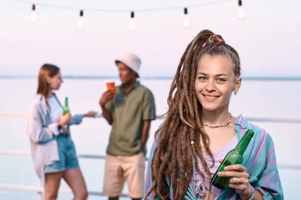 Feliz joven hembra con botella de cerveza de pie contra la pareja intercultural —  Fotos de Stock