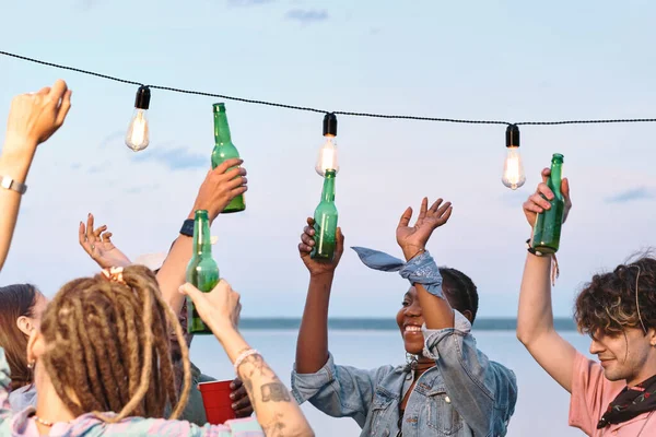 Young ecstatic friends with bottles of beer dancing by waterside — Stock Photo, Image