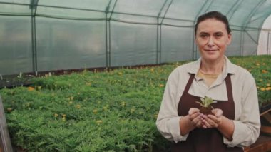 Beyaz önlüklü, orta yaşlı bir kadının orta yavaş portresi. Elinde serada duran küçük yeşil fidanla kameraya poz veriyor.