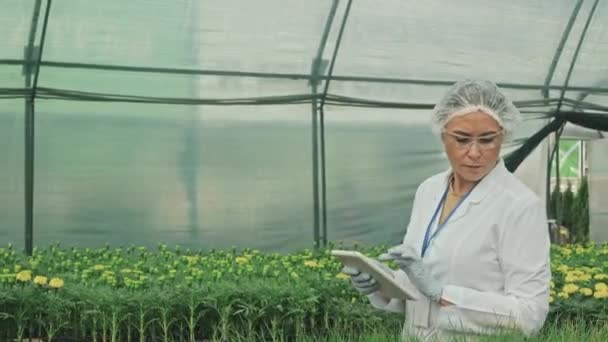 跟踪穿着实验室外套和安全眼镜的女性农业监察员分析植物和花卉生长的照片 并在温室行走时在数字平板上记录 — 图库视频影像