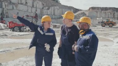 Üniformalı kadın yöneticinin orta boy fotoğrafı, güvenlik gözlükleri ve taş ocağındaki erkek işçilerle konuşan bir şapka. Arka planda çalışan makineler