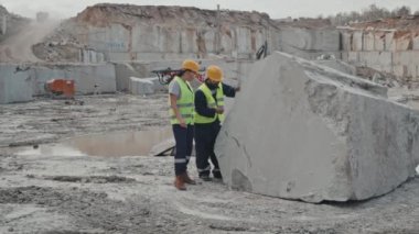 Yelekli kadın ve erkek yöneticilerin yavaş yavaş takibi, güvenlik gözlükleri ve taş ocağındaki büyük çiğ granit taşları teftiş eden sert şapkalar.