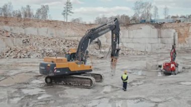 Üniformalı ve kasklı bir erkek işçinin granit ocağındaki sondaj kulesinin yanından geçerken geniş açılı görüntüsü.