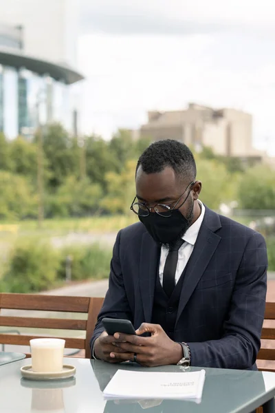 Młody afrykański biznesmen w garniturze i czarnej masce smsowanie przez stół — Zdjęcie stockowe