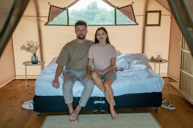 Çift kişilik yatakta oturan, gündelik giyimli, sevgi dolu genç bir çift.