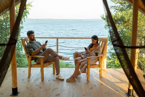 Akıllı telefonları olan genç bir çift glamping evinin terasında oturuyor. — Stok fotoğraf