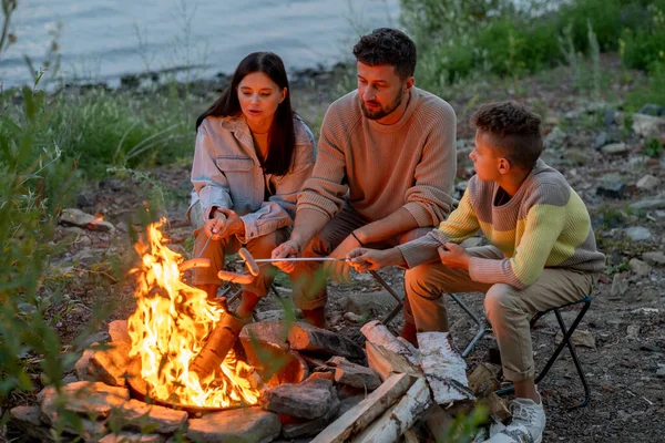 Trzech młodych turystow smaży kiełbaski na ognisku. — Zdjęcie stockowe