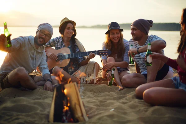 Vrienden met dranken en gitaar door kampvuur — Stockfoto