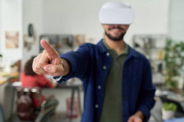 Sanal gerçeklik kulaklığı takan genç bir adam dijital arayüzle etkileşime geçiyor, kollarını uzatmış ve parmağını uzatmış bir şekilde modern çalışma alanında duruyor.