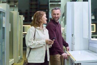 Beyaz bir kadın elinde akıllı bir telefonla mağazada mutfak aletlerini ölçen beyaz bir adamla konuşuyor. İkisi de yan yana duruyor ve gülümsüyor.