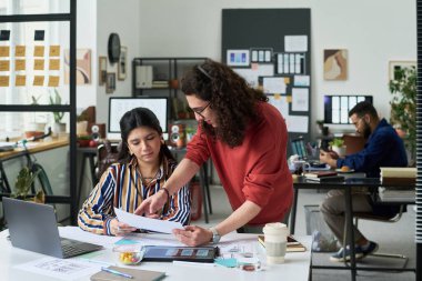 Genç beyaz adam modern ofisteki genç İspanyol kadına belgeleri açıklarken, beyaz adam arka planda dizüstü bilgisayar ve akıllı telefon kullanarak çalışıyor.