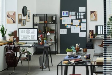 Modern ofis çalışma alanı bilgisayarları, dijital cihazları, bitkileri ve yaratıcı materyalleri gösteren boş masaları, kablolar ve notlarla kaplı duvarı, doğal gün ışığı aydınlatma sahnesini gösteriyor