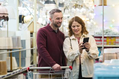 Kafkasyalı genç bir adam aydınlatma mağazasında birlikte alışveriş yaparken elinde akıllı telefon tutan beyaz bir kadının yanında duruyor. Hem gülümsüyor hem de ekranlara bakıyor.