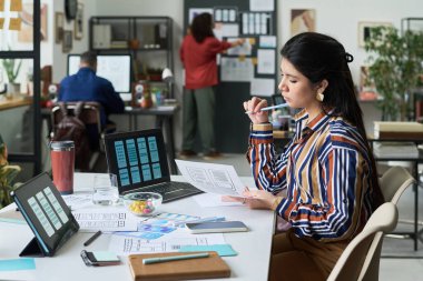 Genç Latin kadın masadaki kablo çizimleri ve dijital düzeni analiz ediyor, ağza yakın bir kalem tutuyor, modern ofiste bilgisayar ve tabletle çalışıyor ve arka planda iş arkadaşlarıyla işbirliği yapıyor.