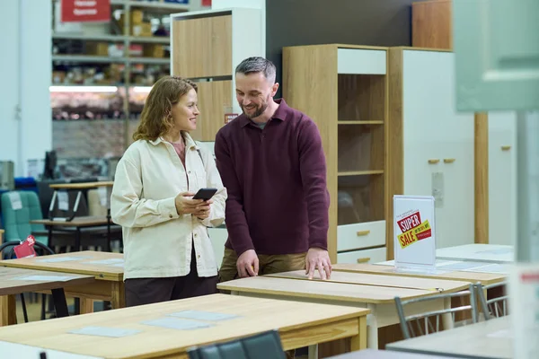 Kafkasyalı bir kadın elinde akıllı telefonla mobilya dükkanında masaların yanında durmuş birbirine gülümseyen ve bakan beyaz bir adamla konuşuyor.