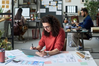 Masada tasarım projesi üzerinde çalışırken Stylus ile dijital tablet kullanan genç beyaz bir adam, modern ofis arka planında işbirliği yapan iki genç iş arkadaşı,