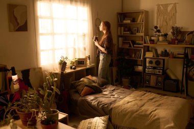 Kafkasyalı genç bir kadın cam kenarında durmuş, ev bitkileri, kitaplıklar ve kişisel eşyalarla dolu rahat bir yatak odasına bakıyor, içeri güneş ışığı giriyor.