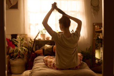 Kafkasyalı genç bir kadın, güneşli yatak odasında saçlarını bağlıyor, kameraya bakıyor, ev bitkileri ve kişisel eşyalarla çevrili, günlük ev ortamında sabah rutini.