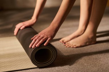 Genç bir kadın iki eliyle yoga minderini yuvarlıyor, halı kaplı zeminde çıplak ayakla duruyor, elleri ve ayakları görünür halde duruyor, antrenmana hazırlanıyor ya da bitiriyor.