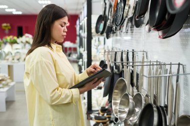 Mutfak gereçleri mağazasında dikilirken dijital tablet kullanan kadın, yemek takımları seçimi ve karşılaştırma seçenekleri, satın almak için uygun kızartma tavası seçmeye odaklandı