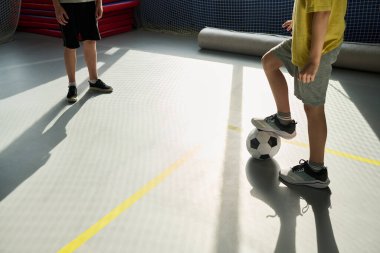 Kapalı alanda futbol oynayan iki çocuk, futbol topuna basan beyaz bir çocuk, spor sahasında hem şort hem de spor ayakkabısı giyen başka bir çocuk ona bakıyor.