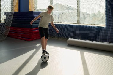 Kafkasyalı bir çocuk futbol topunu iç sahada dengeliyor, spor paspasları ve ağlı pencerelerin yanında duruyor, gündüz antrenmanı sırasında top kontrolü alıştırması yapıyor.
