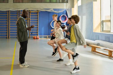 Çok ırklı gençlerden oluşan bir gruba spor salonunda ısınma egzersizleri yaptıran siyahi bir adam. Koordineli hareketlerde dizlerini kaldıran gençler. Arka planda görünen spor ekipmanları.