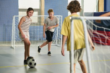 Bir grup Kafkas ve çok ırklı çocuk içeride futbol oynuyor, üç çocuk kale ağlarının yanında aktif olarak hareket ediyor, bir kız top sürüyor, iki erkek de maç sırasında savunma yapıyor.