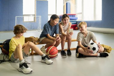 Beden eğitimi bankında oturan dört çocuk, iki kız ve iki erkek, çok ırklı, akıllı telefonla etkileşime geçen, basketbol ve futbol topu tutan, spor aktivitelerinden sonra rahatlayan