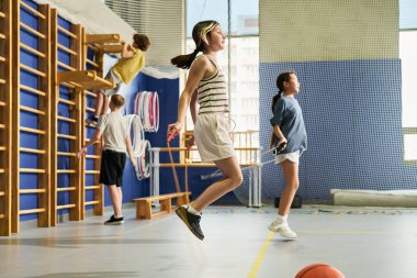 Spor salonunda egzersiz yapan bir grup etnik kökenli genç, ön planda atlayan beyaz bir kız, arkada yürüyen Asyalı bir kız, duvarlara tırmanan bir oğlan, yakınlarda duran başka bir erkek.