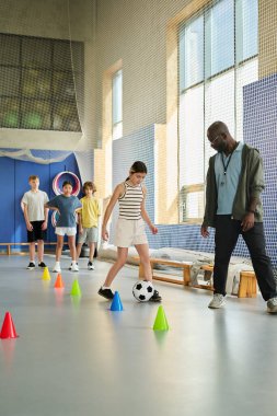 Beyaz bir kız futbol topunu külahlara sürerken siyahi bir adam denetliyor, bir grup çocuk arkada sırada bekliyor, kapalı spor salonu ayarları, beden eğitimi aktiviteleri...
