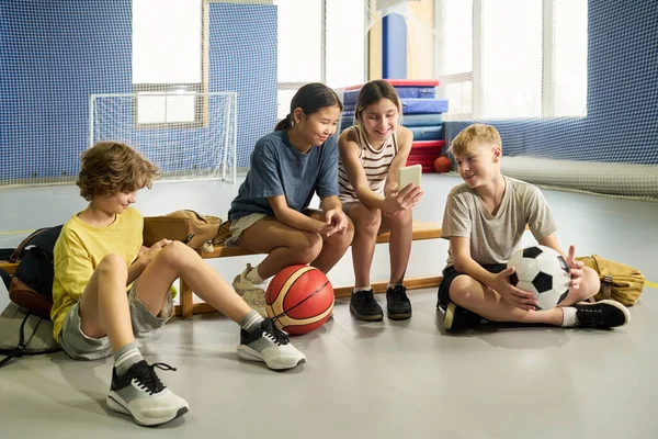 Beden eğitimi bankında oturan dört çocuk, iki kız ve iki erkek, çok ırklı, akıllı telefonla etkileşime geçen, basketbol ve futbol topu tutan, spor aktivitelerinden sonra rahatlayan