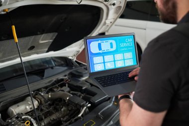 Araba servis arayüzü olan dizüstü bilgisayar kullanan beyaz bir genç adam açık araba kaputunun yanında durup atölyede araç bakımı yapıyor.