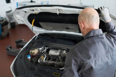 Orta yaşlı beyaz bir adam atölyedeki araba kompartımanını teftiş ediyor, kameraya arkası dönük duruyor, iş eldivenleri giyiyor, kaputu açık tutuyor, araç bakımına odaklanıyor.