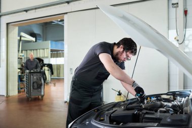 Otomobil tamirhanesinde araba motoru tamir eden, iş eldiveni ve tulum giyen, orta yaşlı, beyaz bir adam arka planda dikilirken mekanik işe odaklanmış beyaz bir adam.