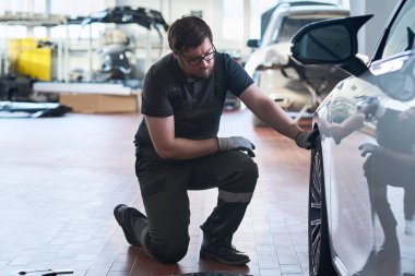 Beyaz, yetişkin bir adam araba tamirhanesinde araba teftişi tekerinin yanında diz çöküyor, iş eldiveni ve tulum giyiyor, profesyonel garaj ortamında bakım işine odaklanıyor.