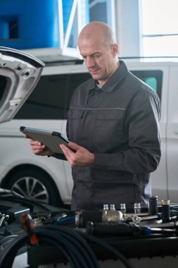 Orta yaşlı beyaz adam araba motorlarını kontrol ederken dijital tablet kullanarak oto tamirhanesinde duruyor, profesyonel çevrede teşhis ve bakım görevlerine odaklanıyor
