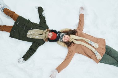 Kafkasyalı ergen çocuk ve annesi kar melekleri yaparak, gülümseyip yukarı bakıyorlar, kış kıyafetleri ve eşarpları görünür, açık hava kış aktiviteleri