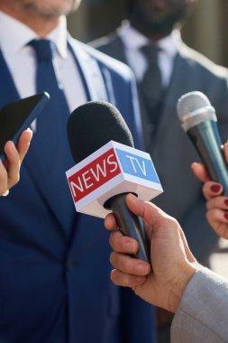 Beyaz, orta yaşlı, takım elbiseli bir adam basın toplantısı sırasında elinde mikrofonla gazetecilere konuşuyor. Ön planda görünen gazetecilerin elleri, medya etkileşimine odaklanıyor.