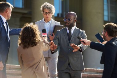 Orta yaşlı, beyaz takım elbiseli, elinde klasör tutan beyaz bir adam. Takım elbiseli genç siyahi bir adam jest yaparken durdu. Dışarıda mikrofonları ve kameraları olan bir grup gazeteci.
