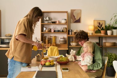 Kafkasyalı genç bir kadın mutfak masasında beyaz bir çocuğu kucağında tutarken meyve suyu dökerken, aile kahvaltıda meyve ve doldurulmuş oyuncakla etkileşime geçiyor.