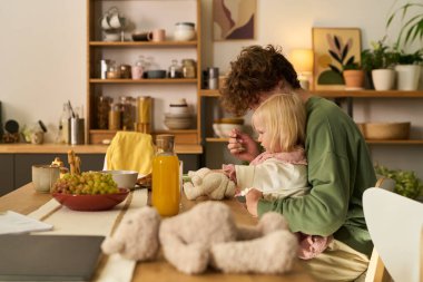 Kafkasyalı ergen, beyaz kızı yemek masasında birlikte otururken besliyor, yemeğini paylaşıyor ve sıcak mutfakta vakit geçiriyor, masada içi doldurulmuş oyuncak ve meyve suyu var.