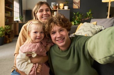 Kafkasyalı bir kadının beyaz bir kızı kucağında tutarken beyaz bir gencin birlikte selfie çektiği, gülümseyen ve rahat oturma odasında kameraya bakan portresi.