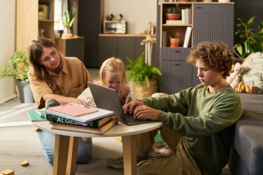 Beyaz, genç, yetişkin bir kadın, beyaz bir kıza yardım ederken, beyaz bir erkek, oturma odasında dizüstü bilgisayar kullanıyor. Kitaplar ve oyuncaklar etrafa saçılmış.